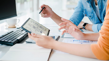 mensen aan bureau met ipad. In beeld zijn alleen de armen, handen en bovendeel van de personen.